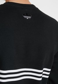 Svart sweatshirt med vita horisontella ränder, prydd med en liten broderad svart logotyp på övre ryggen. Tillverkad av bomull, grundläggande rund hals.