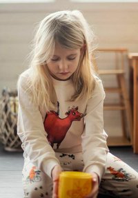 Lapsi, jolla on yllään beige collegepaita, jossa on suuri punainen lehmän grafiikka, ja valkoiset housut, joissa on eläinkuvioita, istuu ja pitää keltaista mukia käsissään.