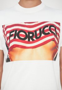 Άτομο που φοράει λευκό μπλουζάκι με σχέδιο σε κόκκινες και λευκές ρίγες και τη λέξη "FIORUCCI" με έντονους μαύρους χαρακτήρες.