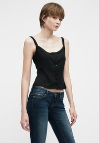 Joven con cabello corto castaño, vistiendo una camiseta sin mangas negra con botones y jeans azul oscuro, de pie contra un fondo blanco liso.