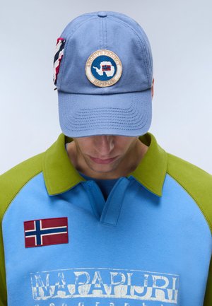 Mies, jolla on sinivihreä kauluksellinen collegepaita, jossa on Norjan lipun merkki, ja vaaleansininen lippalakki, jossa on pyöreä logo ja lipun yksityiskohta sivulla.