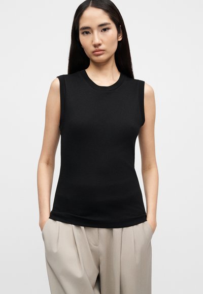 Γυναίκα με μακριά σκουρόχρωμα μαλλιά, που φοράει μαύρο αμάνικο τοπ και μπεζ πλισέ παντελόνι, στέκεται με τα χέρια στις τσέπες μπροστά σε λευκό φόντο.