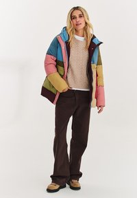 Vícebarevná péřová bunda se vzorem v modré, růžové, zelené a vínové barvě; má kapuci, zip a boční kapsy; nošena přes béžový svetr.