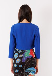 Giacca blu tagliata, realizzata in tessuto morbido, con maniche a tre quarti e scollatura rotonda, abbinata a una gonna colorata a fantasia.