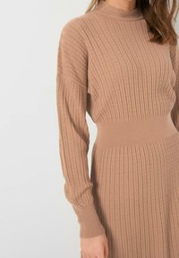 Stickad klänning i ljusbeige färg med hög krage, ribbad textur och långa, figurnära ärmar. Dragen midja ger extra form.