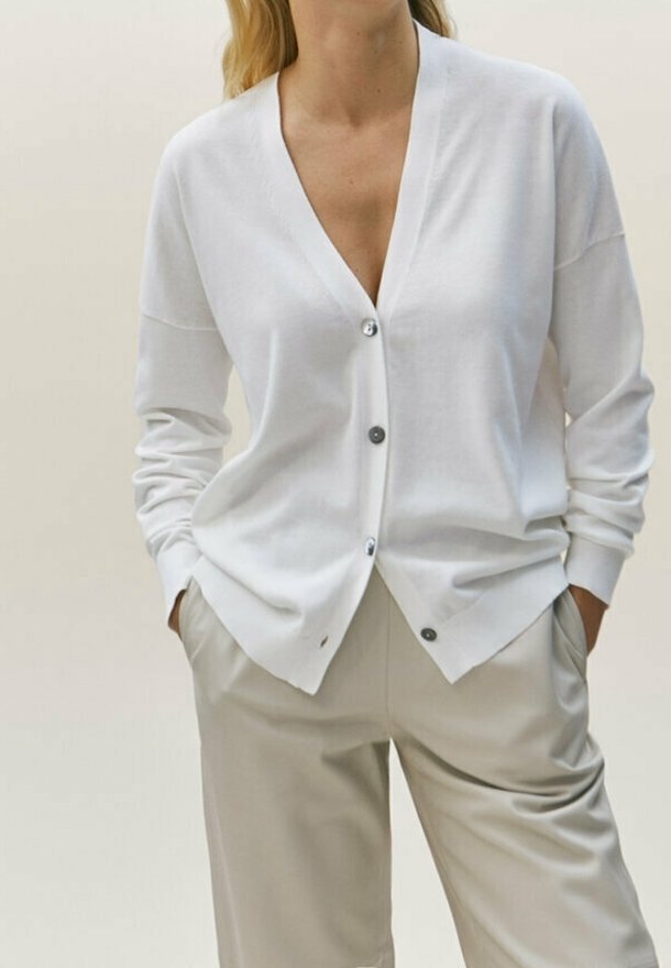 White cardigan with a v-neck, long sleeves, and gray buttons, paired with beige pants. Soft texture and relaxed fit.