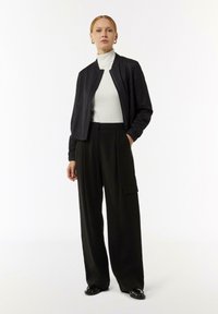 Zwarte bomberjack met een ritssluiting, gedragen over een witte turtleneck. Gecombineerd met losse, high-waisted zwarte cargobroeken en glanzende zwarte schoenen.