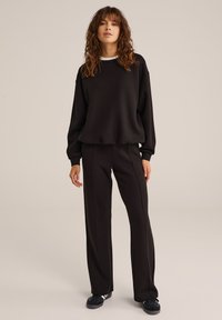 Felpa nera con scollatura a girocollo e spalle cadenti, abbinata a pantaloni neri con cuciture verticali. Il materiale appare morbido e texturizzato.