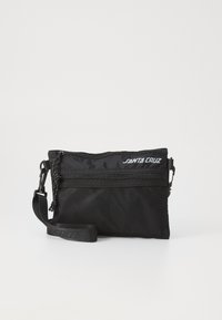 Zwarte stoffen schoudertas met gaasdetails, ritsluiting, verstelbare schouderband en "Santa Cruz" logo aan de voorkant. Compact rechthoekig van vorm.
