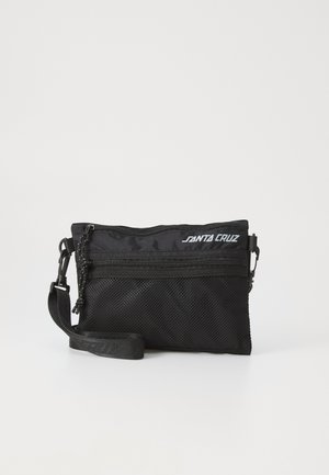 Sac à épaule en tissu noir avec des détails en maille, fermeture éclair, sangle ajustable et logo "Santa Cruz" à l'avant. Forme rectangulaire compacte.