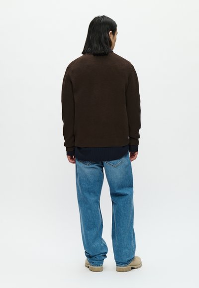 Pulôver castanho tricotado com uma textura canelada, combinado com jeans largos de cor azul clara. O conjunto apresenta uma camisa escura em camadas por baixo.