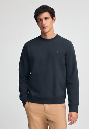 Granatowy sweter z miękkiego materiału, z okrągłym dekoltem, długimi rękawami i małym czerwonym logo po lewej stronie klatki piersiowej. Prosty design, bez wzorów.