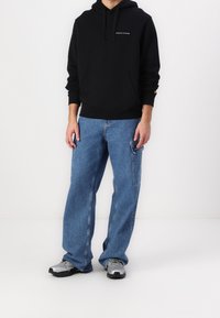 Personne portant un hoodie noir avec le logo "Lyle & Scott", un jean bleu ample avec des poches latérales, et des baskets gris argentés se tenant sur un sol blanc.