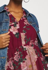 Vestido floral en burdeos con estampados en rosa y verde, con botones y cintura fruncida, llevado debajo de una chaqueta de mezclilla azul.