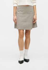 Άτομο που φοράει φούστα σε μπεζ και μαύρο καρό, λευκές κάλτσες και μαύρα-άσπρα παπούτσια, στέκεται μπροστά σε ένα απλό λευκό φόντο.