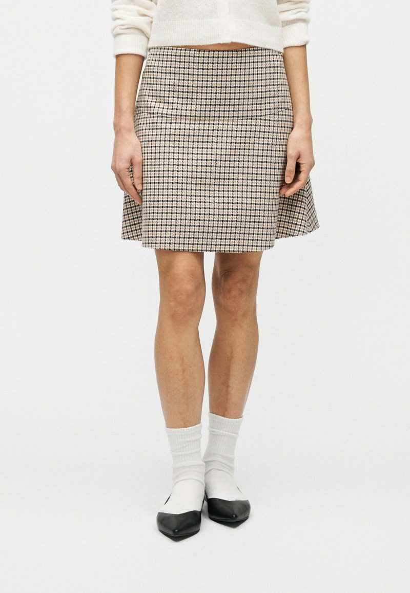 Άτομο που φοράει φούστα σε μπεζ και μαύρο καρό, λευκές κάλτσες και μαύρα-άσπρα παπούτσια, στέκεται μπροστά σε ένα απλό λευκό φόντο.