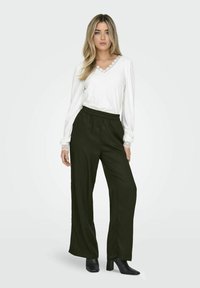 Hvid langærmet bluse med blondekant og V-udskæring, kombineret med mørkegrønne vide bukser og sorte hælede ankelstøvler.