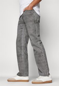 Šedé džínové kalhoty s volným střihem, s kontrastním prošíváním a bočními kapsami, v kombinaci s bílými teniskami a jednoduchým bílým tričkem.