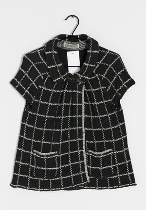 Veste noire à manches courtes avec motif à carreaux blancs, large col, fermeture à boutons sur le devant, deux poches, suspendue sur un cintre noir sur fond blanc.