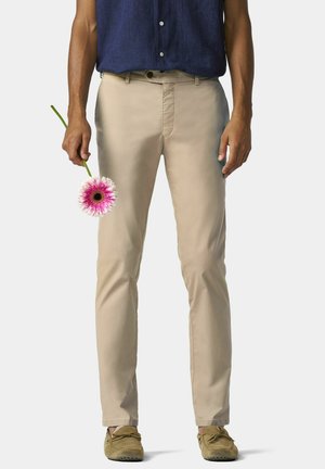 Lichtbeige katoenen broek met een rechte pasvorm, voorzakken en een subtiele glans. Een hand houdt een roze bloem vast voor contrast.