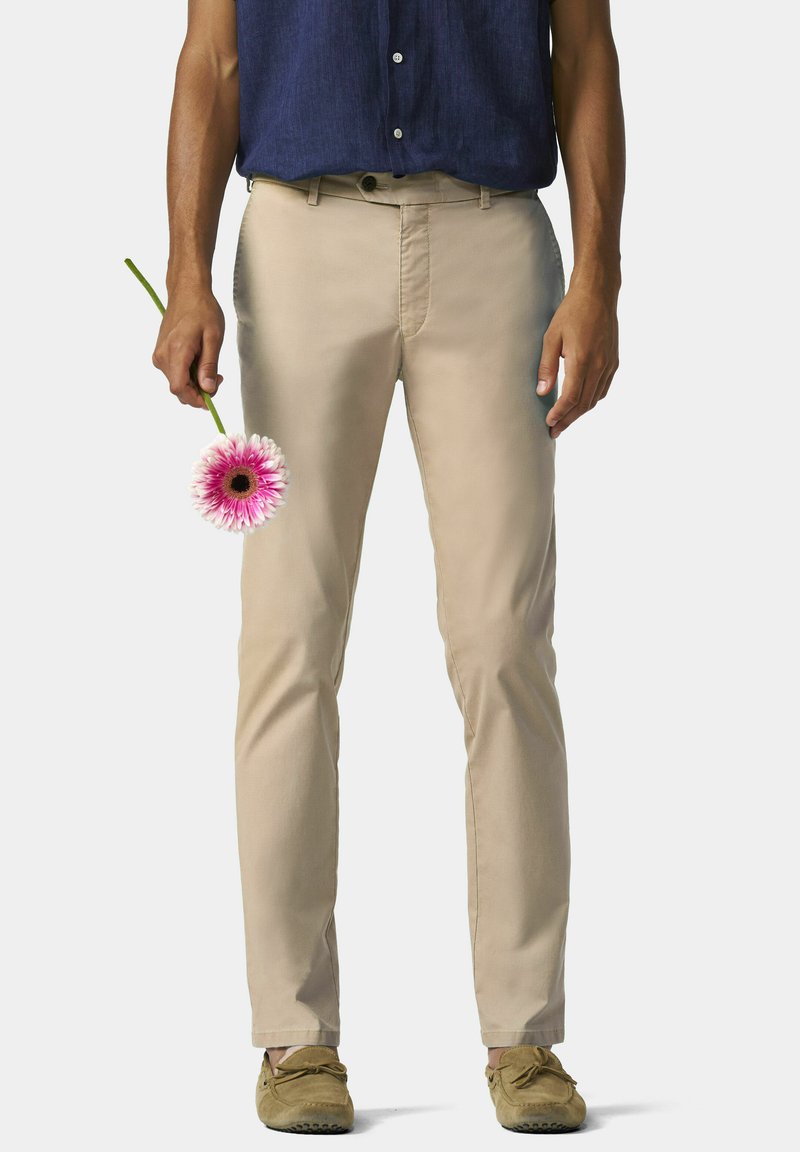Helle beige Baumwollhosen mit geradem Schnitt, Fronttaschen und einem dezenten Glanz. Eine Hand hält eine rosa Blume zur Kontrastierung.