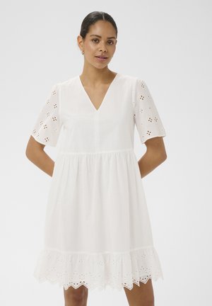 Žena v bielych šatách s výstrihom do V, s kvetinovými dierkami na rukávoch a priečnym okrajom, stojaca s rukami za chrbtom.