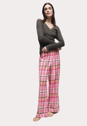 Lange, rosa karierte Schlafanzughosen mit schwarz-weißen Akzenten, kombiniert mit einem figurbetonten, dunklen Langarmoberteil mit V-Ausschnitt und Knopfdetails.