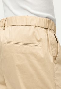 Gros plan d'un pantalon beige à taille élastique avec une poche passepoilée arrière et une poche sur la couture latérale, porté avec un haut blanc côtelé.