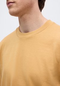 Άνδρας που φοράει απλό μουσταρδί κίτρινο t-shirt με στρογγυλή λαιμόκοψη, εμφανίζοντας τον λαιμό και το κάτω μέρος του προσώπου με ελαφρύ τριχωτό υπόβαθρο, σε απλό φόντο.