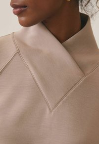 Jasno-beżowa bluzka z wysokim kołnierzem, charakteryzująca się strukturalnym krojem, miękką teksturą oraz widocznymi detalami stębnowania na kołnierzu i szwach.