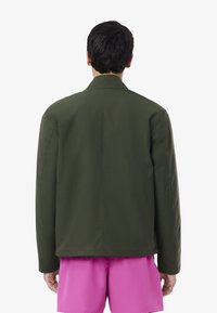 Giacca leggera verde oliva con colletto, vestibilità ampia e cuciture minime. Indossata sopra pantaloni corti rosa brillante, mostrando una texture liscia.