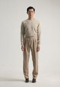 Camisa beige de manga larga con pequeño logo, combinada con pantalones beige de talle alto con pliegues y zapatos oscuros, de pie sobre un fondo neutro.