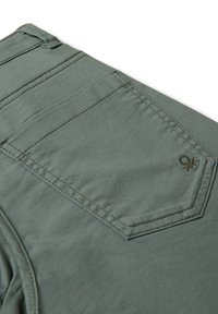 Acerca del bolsillo trasero de unos pantalones gris-verde, con costuras visibles y un pequeño logo bordado cerca del borde del bolsillo.