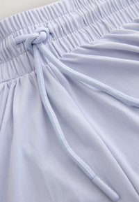 Ceinture élastique avec un cordon blanc noué sur un tissu lavande clair, montrant des sections froncées et lisses du vêtement.