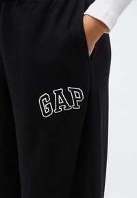 Άτομο με το χέρι στην τσέπη, φοράει μαύρο φούτερ παντελόνι με λευκό λογότυπο «GAP» στο μηρό και λευκό μακρυμάνικο μπλουζάκι.