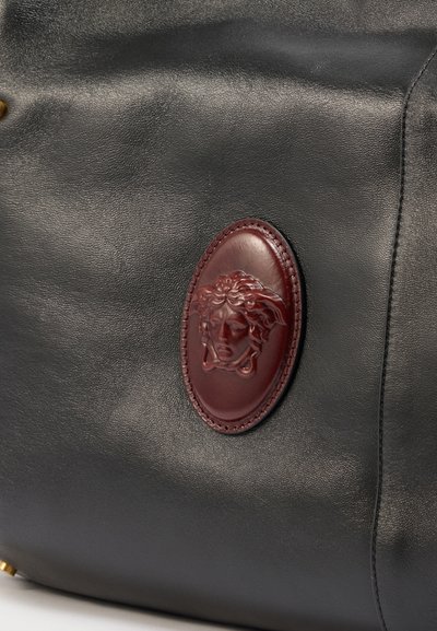 Sort lædertaske med et syet ovalt bordeaux medaljon, der har et forhøjet detaljeret ansigtsemblem i midten.