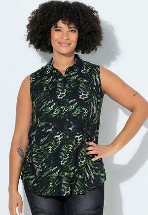 Kvinde med krøllet hår, der smiler, iført en sort ærmeløs knap-op bluse med grøn og hvid dyreprint.