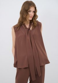 Bruine mouwloze blouse van lichte stof, met een centrale strik en plooien bij de halslijn, wat een vloeiende silhoutte biedt.