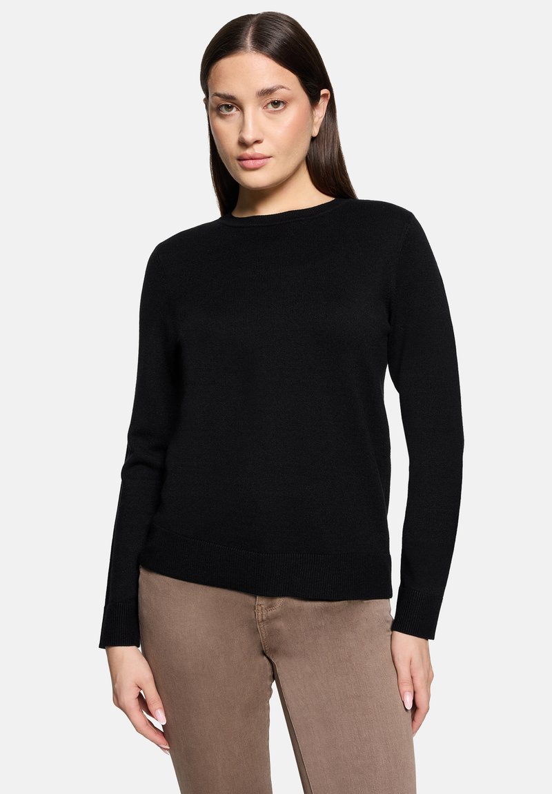 Jeune femme aux cheveux bruns raides portant un pull noir à manches longues uni et un pantalon marron clair, debout devant un fond blanc.