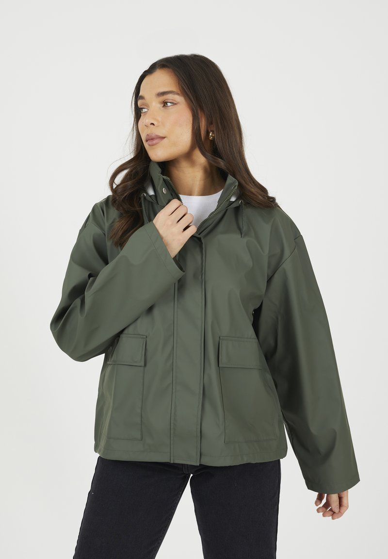 Veste imperméable verte avec capuche, poches avant et coupe décontractée. Sa texture lisse et son design minimaliste mettent en valeur sa fonctionnalité.