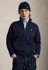 Ung mann med marineblå glidelåsjakke, lyseblå skjorte, mørke jeans og marineblå strikket lue, med hendene i lommer mot nøytral bakgrunn.