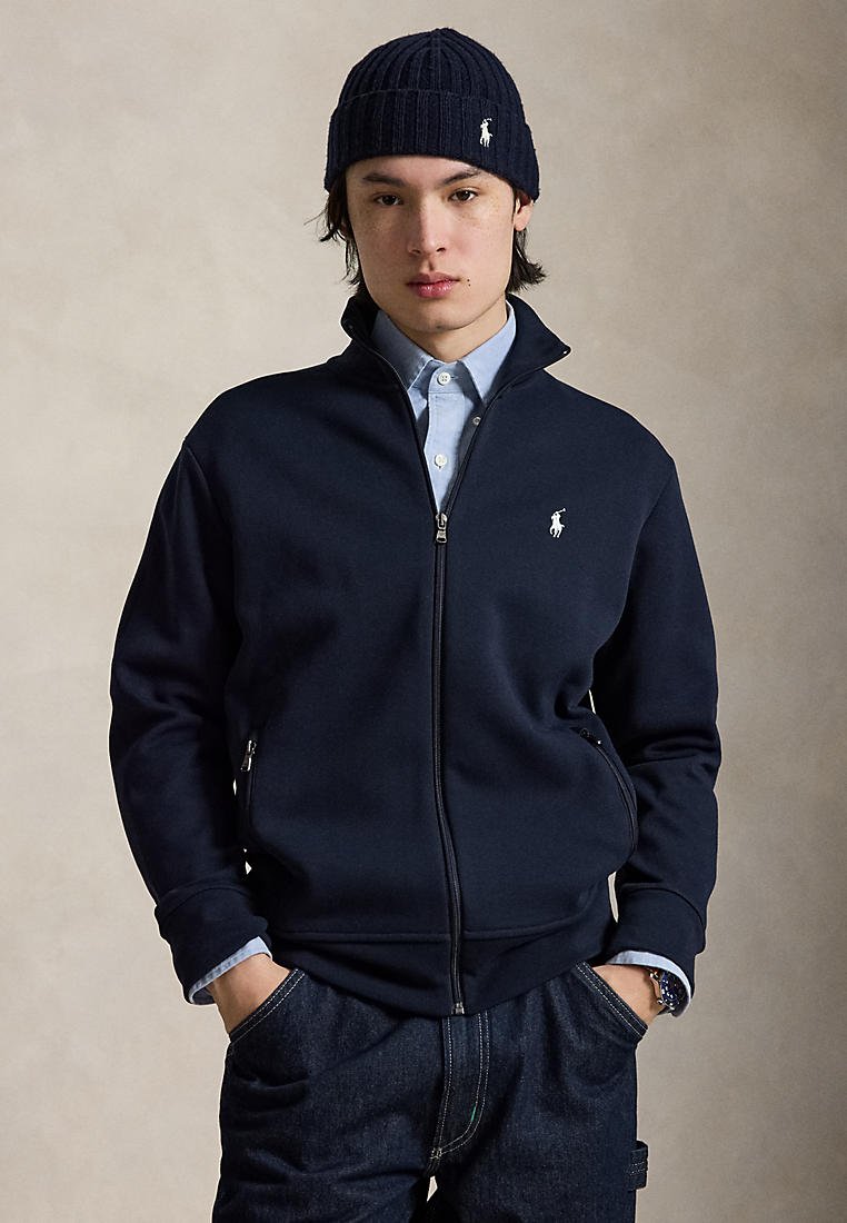 Jeune homme portant une veste zippée bleu marine, une chemise bleu clair, un jean foncé et un bonnet en tricot bleu marine, les mains dans les poches, sur un fond neutre.