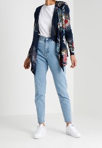 Marinblommig cardigan med långa ärmar, öppen fram och asymmetrisk fåll, i kombination med ljusblå hög midja jeans och vita sneakers.