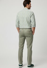 Camisa de cuadros verde de hombre, pantalones caqui y zapatillas verdes, con las mangas arremangadas y bolsillos visibles en la parte trasera de los pantalones.