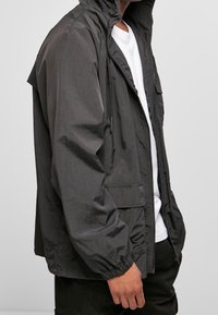 Black, lightweight jacket with a hood, zip front, elastic cuffs, and a chest pocket; features a smooth, slightly reflective texture.