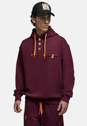 Jeune homme portant un sweat à capuche bordeaux avec des cordons orange et un pantalon assorti, accompagné d'une casquette noire avec un patch coloré.