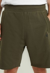 Pantaloni sportivi verde scuro con una texture liscia, vita elastica e tasche laterali. Presentano un piccolo logo nero sulla coscia sinistra.