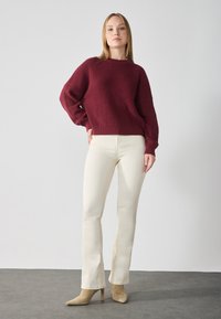 Burgundy ribbed knit sweater with wide sleeves paired with cream bootcut pants and beige high-heel ankle boots on a gray floor.