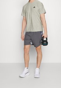 Grå atletisk T-shirt med en svart logotyp, tillsammans med grå shorts med ett mönster i blå accenter. Personen håller en svart kettlebell.