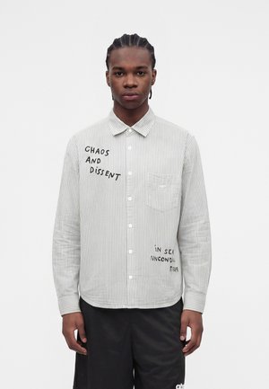 Jonge man met gevlochten haar die een wit-grijs gestreept overhemd met knopen draagt met zwarte tekst "Chaos and Dissent" op de borst, zwarte broek.